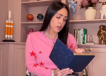 Una mujer con suéter rosa escribe en un cuaderno azul frente a una estantería con libros, maquetas de cohetes y figuras de astronautas