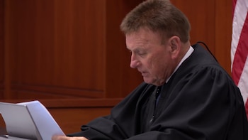 Un hombre de mediana edad con toga judicial negra mira documentos sobre una mesa en una sala de tribunal con paredes de madera y bandera estadounidense