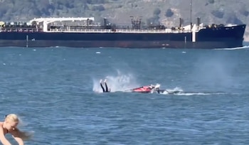 Un kayak rojo volcado en el agua con las piernas de una persona sobresaliendo, rodeado de salpicaduras. Un gran barco oscuro se ve navegando en el fondo