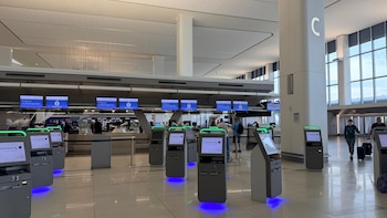 Interior de aeropuerto moderno con kioscos de auto check-in de American Airlines, pantallas digitales y viajeros. Se aprecian grandes ventanas al fondo. Aeropuerto de La Guardia de Nueva York