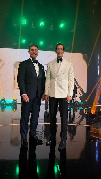 Dos hombres en esmoquin posan de pie en un escenario iluminado con luces verdes y naranjas; uno lleva un bigote y esmoquin negro, el otro un esmoquin blanco