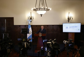 Hombre con traje y gafas habla en un podio. Bandera de Guatemala a la izquierda, pantalla digital con documentos a la derecha. Fondo con paneles de madera