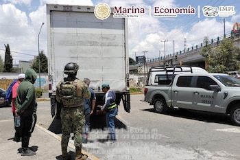 Un militar con uniforme de camuflaje observa a varios hombres junto a un camión de carga blanco estacionado en la calle junto a una camioneta