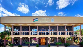 Fachada de una embajada con letrero "Embajada de Palestina", bandera y escudo de Palestina, balcones, columnas y un cartel de "Bienvenidos". Cielo azul con nubes