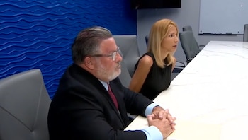 Un hombre con gafas y traje formal junto a una mujer de cabello rubio sentados a una larga mesa blanca en una sala con pared azul