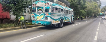 Bus colorido estacionado en carretera, varios agentes de tránsito en chalecos amarillos, camión rojo, pickup blanco, árboles y cinta amarilla en el suelo