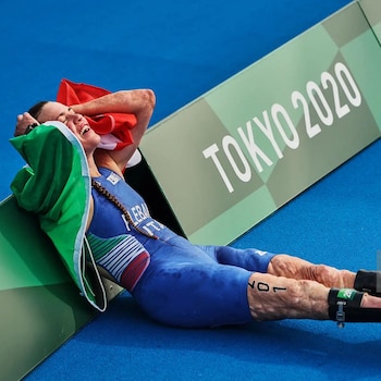 Una mujer vestida con un traje azul e insignias de Italia yace en el suelo azul y sostiene una bandera italiana junto a un cartel de Tokio 2020