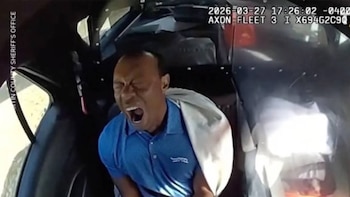 Primer plano de Tiger Woods dentro de un vehículo oscuro, con una camisa polo azul, la boca abierta y los ojos cerrados, mostrando angustia