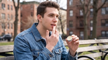 Hombre joven aplica bálsamo labial en un envase metálico pequeño con su dedo en un parque, con edificios de ladrillo y árboles de fondo.