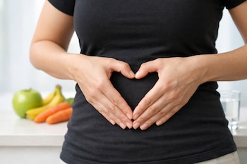 Manos sobre un abdomen forman un corazón. Una manzana verde, plátanos, zanahorias y un vaso de agua son visibles en el fondo desenfocado.