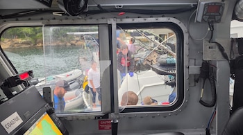 Interior de una cabina de barco con ventanales que muestran personas, botes auxiliares y redes de pesca en el agua con una costa arbolada.