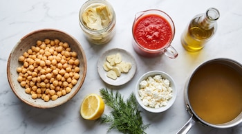 Garbanzos, alcauciles en conserva, tomate triturado, aceite, ajo fileteado, limón, eneldo, queso desmenuzado y caldo sobre una superficie de mármol.