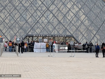 Personas con pancartas frente a la entrada acristalada de la pirámide del Museo del Louvre, detrás de postes delimitadores en la explanada