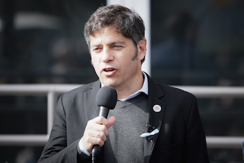 Hombre con cabello oscuro y traje hablando a un micrófono. Lleva un suéter gris, una chaqueta negra y pines en la solapa izquierda. Tiene un anillo en la mano