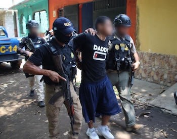 Dos agentes de policía armados escoltan a un hombre con camiseta negra y pantalón corto azul por una calle
