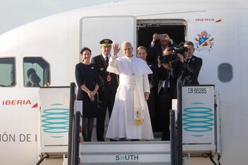 El Papa León XIV vestido de blanco saluda desde la puerta abierta de un avión blanco de Iberia, rodeado de personal de cabina y fotógrafos