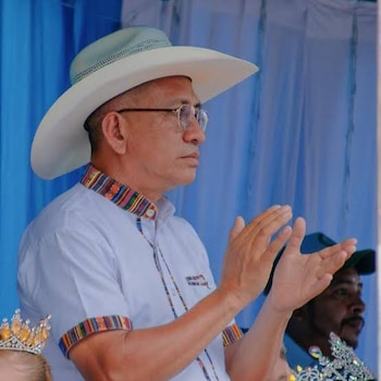 Hombre de perfil con sombrero vaquero claro, gafas, camisa blanca con bordes de diseño étnico, aplaudiendo. Fondo azul y dos coronas