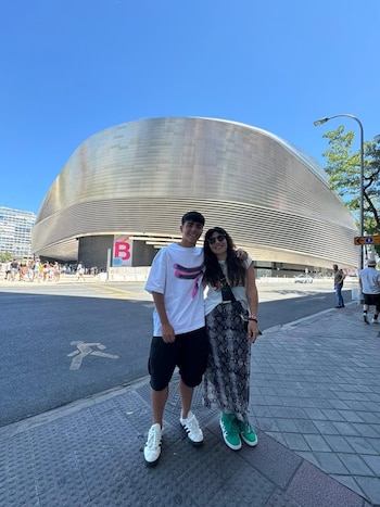 Una mujer y un joven sonríen a la cámara, de pie en una acera. Detrás de ellos, un gran edificio moderno y curvo de fachada metálica bajo un cielo azul despejado