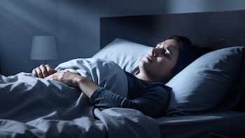 Una mujer acostada en una cama con mantas grises expresa malestar con los ojos cerrados en un dormitorio a oscuras