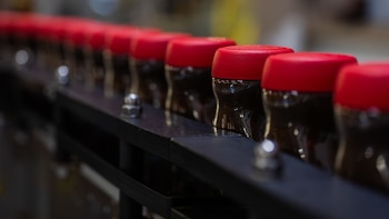 Una hilera de frascos plásticos oscuros con tapas rojas brillantes sobre una banda transportadora metálica con fondo borroso