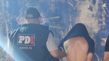 Un agente de PDI Santa Fe, de espaldas, sujeta a un hombre con la camiseta sobre la cabeza frente a una pared azul deteriorada
