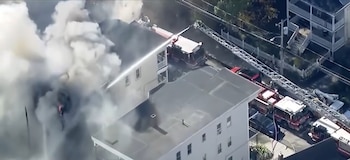 Vista aérea de camiones de bomberos que arrojan agua hacia un edificio residencial del cual sale una nube de humo blanco
