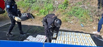 Dos agentes uniformados con chalecos tácticos manejan paquetes rectangulares con logos FIFA y D&G sobre lona negra. Fondo con vegetación y restos de vehículo