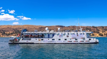 Un barco blanco con la bandera de Perú y distintivos estatales navega en un lago frente a colinas bajo un cielo azul.
