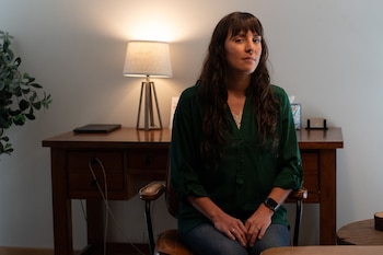 Mujer de cabello castaño oscuro sentada en una silla de oficina de cuero, con blusa verde y jeans, mirando directamente a la cámara. Detrás hay un escritorio de madera con una lámpara encendida y una planta a la izquierda