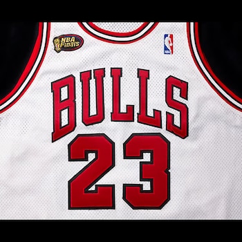 Camiseta blanca de baloncesto con las letras rojas BULLS y el número 23, con bordes negros. Incluye parches de las Finales NBA y el logo de la NBA