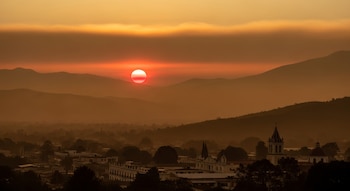 El sol se oculta entre montañas sobre un pueblo con casas, árboles y una torre de iglesia bajo un cielo anaranjado.