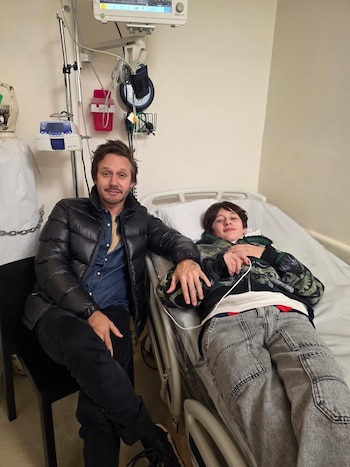Un hombre y un niño en una habitación de hospital. El hombre está sentado en una silla, el niño está recostado en una cama. Hay equipos médicos en la pared