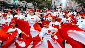 Un grupo de personas, mayormente mujeres y niños, desfilan bailando y llevando trajes rojos y blancos con el texto "I love Bomba" en sus camisetas blancas