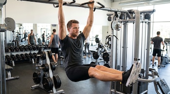 Hombre de tez clara y cabello corto, en camiseta gris y pantalones cortos negros, cuelga de una barra de dominadas con las piernas extendidas, en un gimnasio.