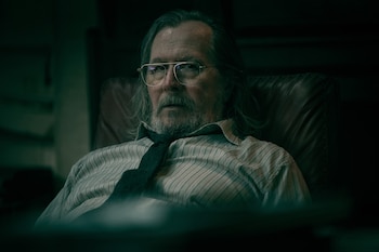 Retrato de un hombre mayor con gafas, barba, cabello canoso, camisa a rayas y corbata floja, sentado en un sillón de cuero