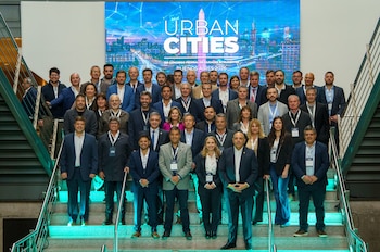 Un grupo de hombres y mujeres en traje posa en las escaleras frente a una pantalla azul que dice Urban Cities