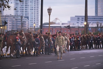 Vista de una calle con soldados uniformados y caballos alineados. Un soldado en camuflaje camina en primer plano. Edificios altos se ven al fondo