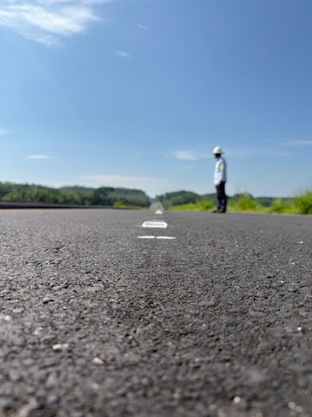 Una vista de cerca de una carretera de asfalto oscuro con marcas viales blancas. En la distancia, una figura borrosa de una persona con chaleco y casco