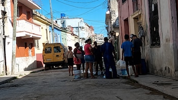 Grupo de personas con cubetas y recipientes en una calle de Cuba, con edificios coloridos y un taxi amarillo bajo un cielo azul