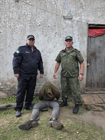 Un policía y un soldado uniformados están de pie junto a una persona sentada en el suelo con la cabeza cubierta, frente a una pared de textura irregular