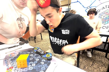 Un joven con antifaz en la frente y camiseta negra mira un cubo Rubik amarillo sobre una alfombrilla y un cronómetro en una mesa