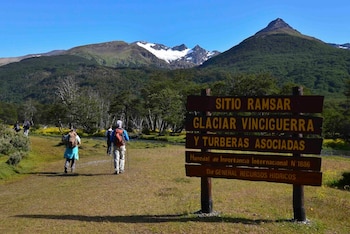 Senderistas en un camino de tierra y pasto junto a un cartel de madera "Sitio Ramsar Glaciar Vinciguerra" con montañas nevadas y bosque. Cielo azul claro