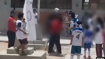 Niños con ropa deportiva y adultos permanecen en el patio de un colegio cerca de una bandera blanca y una imagen difuminada