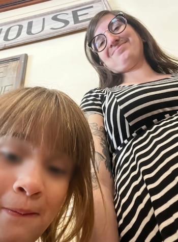 Una mujer con anteojos y vestido a rayas mira hacia la cámara junto a una niña con cabello claro y flequillo