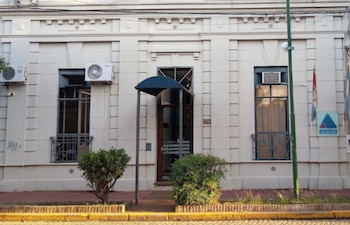 Fachada de edificio blanco con tres ventanas, puerta central con toldo, dos unidades de aire acondicionado, arbustos, cartel 'ACUSACION' y bandera