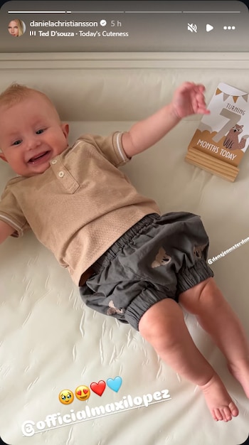 Un bebé rubio con camisa beige y shorts grises con koalas sonríe acostado junto a un cartel que indica siete meses de edad