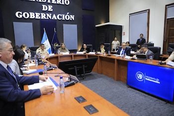 Funcionarios en reunión, Ministerio de Gobernación, Guatemala. Hombres y mujeres en mesa curvada. Logo y nombre del ministerio visible en pantallas y pared