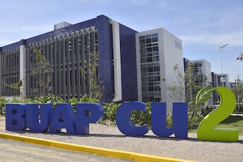 Letrero azul "BUAP CU" y un "2" verde con una hoja en primer plano, frente a edificios universitarios modernos de tonos azules y grises bajo un cielo claro