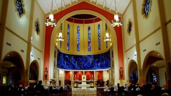 Interior de una iglesia con techos altos, arcos, múltiples vitrales de colores, luminarias colgantes, un altar central y un gran mural azul