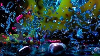 Salpicaduras de líquido azul, fragmentos cristalinos, pequeñas figuras magenta y un fondo desenfocado verde y amarillo